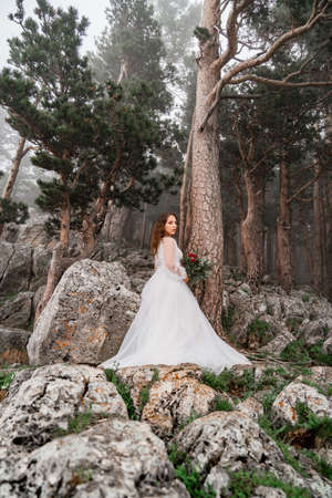 The bride with a bouquet of flowers stands on a rock in the mountain forest.の写真素材