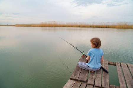 rear view. kid girl sits on a wooden bridge on a river with fishing rod.の写真素材