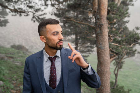 a man with a beard smokes a cigarette standing in the woods by a tree.