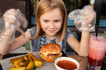 a little girl with gloves eats a burger and potatoes in a summer outdoor cafe.の写真素材