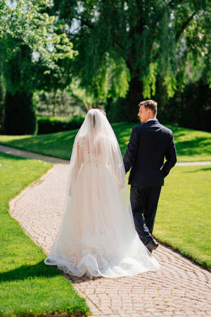the bride and groom walk holding hands along the path in the park.の写真素材