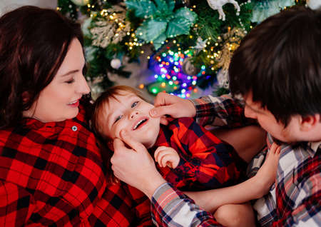 dad, mom and little daughter in red plaid pajamas playing at the Christmas treeの写真素材