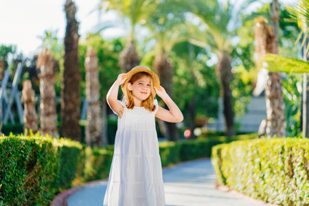 little girl in a white dress and a straw hat on the path in a tropical hotel