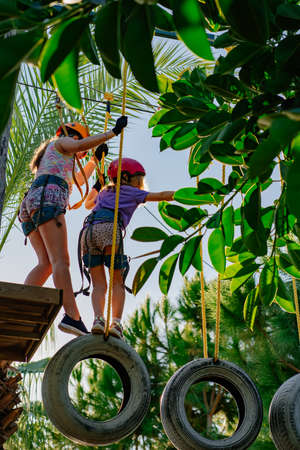 two kids girls in helmet, T-shirt and shorts climbs in rope park at altitude
