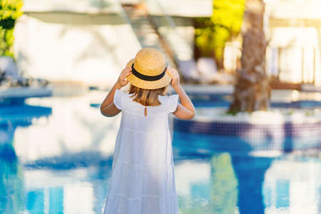 rear view. cute girl in a white dress and straw hat by the pool near the villa.の写真素材