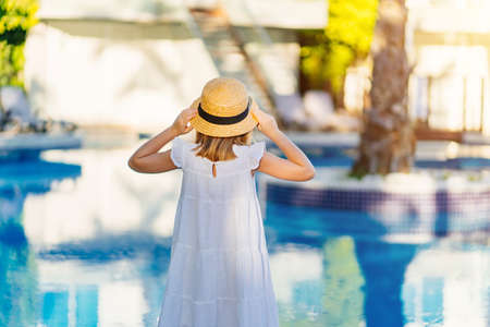 rear view. cute girl in a white dress and straw hat by the pool near the villa.の写真素材