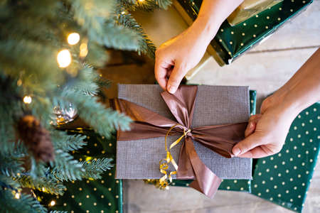 hands tie a brown ribbon on a gray gift box under the christmas treeの写真素材
