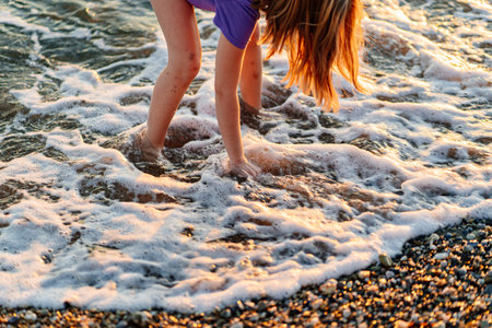 at sunset. legs of girl walking along seashore and throwing stones into waterの写真素材