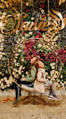 two little girls sit on a swing against a wall of flowers.の写真素材