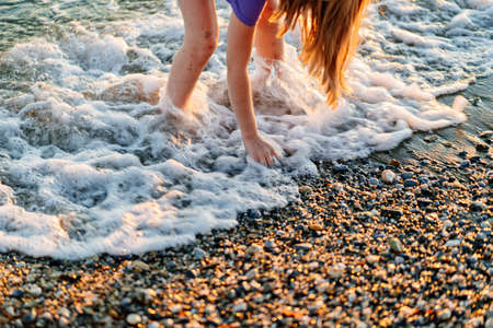 at sunset. legs of girl walking along seashore and throwing stones into water