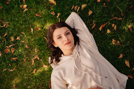 an attractive woman in a hoodie lies on a green lawn with autumn leaves.