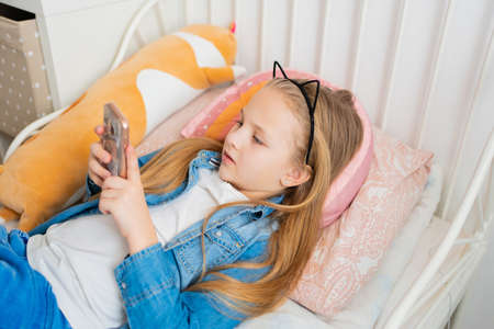 a pretty little girl in a rim with cat ears with a phone in bed.の写真素材