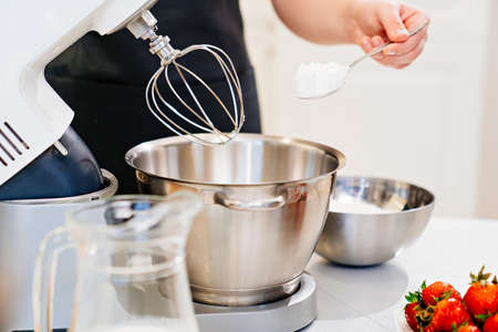 A cook adds a spoonful of flour to prepare dough in a mixer bowl.
