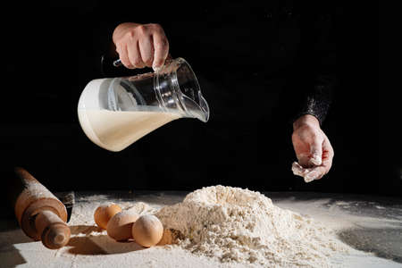 woman pours milk from a jug into flour to knead the dough on black background.の写真素材