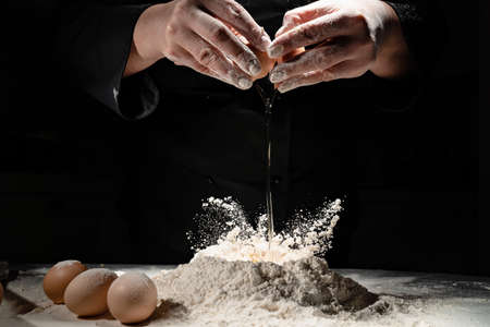 woman breaks the egg into flour to knead the dough on black background.の写真素材