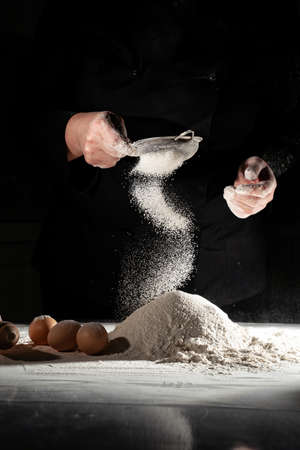 woman sifts flour through sieve onto table with eggs on black background.の写真素材
