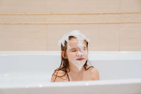 a little cheerful girl with foam on the head washes and plays in the bathroom.