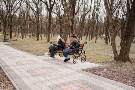 a woman and a girl in black jackets sit on a bench with smartphones.の写真素材