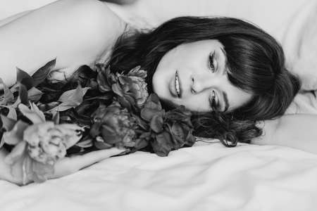 an attractive and sexy young woman with a bouquet of peonies lies in bed.