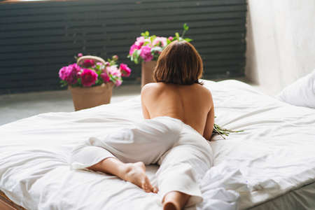 view from back. beautiful topless woman with a bouquet of peonies on the bed.の写真素材