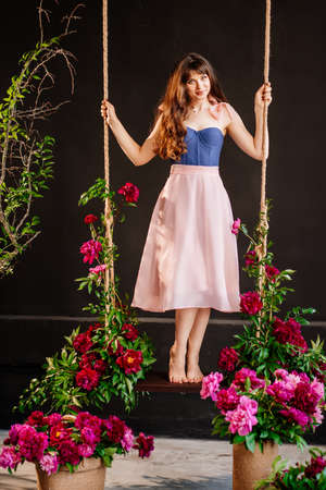 a beautiful young woman in a dress stands on a swing in peony flowers.の写真素材