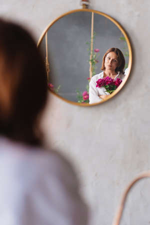 a woman in white with a peonies looks at the reflection in a round mirror.の写真素材