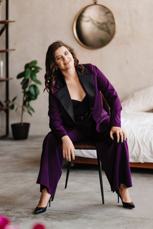 a young brunette woman in a purple pantsuit on a chair in a room with a bed.の写真素材