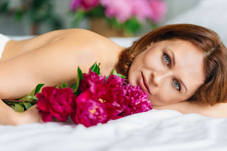 beautiful brunette topless woman with a bouquet of peonies on the bed.の写真素材