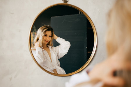 a beautiful blonde in a white shirt looks at her reflection in a round mirror.の写真素材