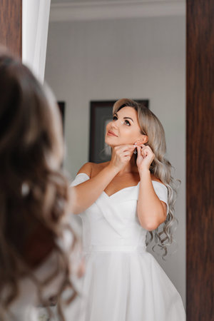 blonde bride in a white dress looks at the reflection and puts on earrings.の写真素材