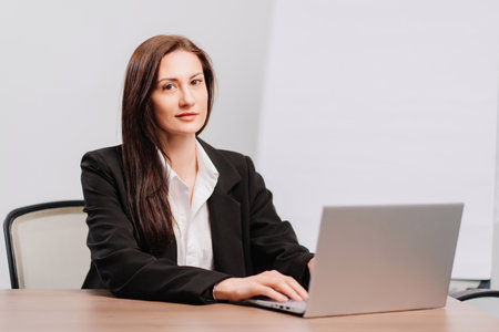brunette woman in black coat in office with meeting board, desktop and laptop