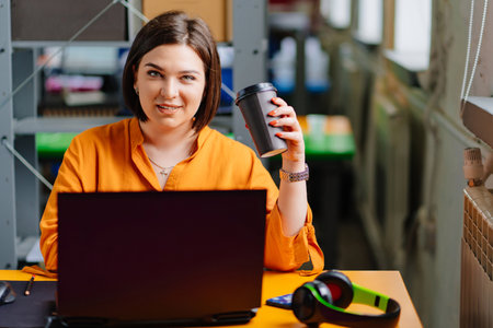 a brunette woman in an orange blouse drinks coffee and works on a laptop