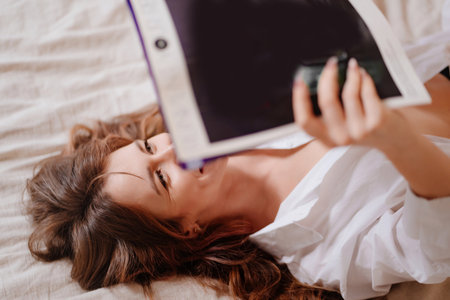 an attractive and happy woman lies in bed and reads a magazine.の写真素材