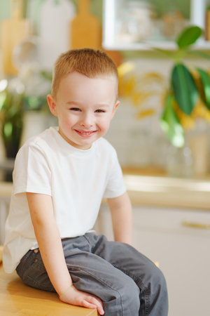 a cheerful cute little boy in a white T-shirt and gray jeans sits on the table.の写真素材