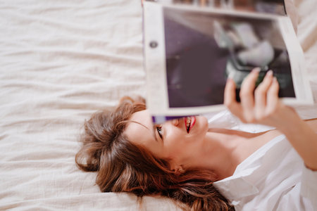 an attractive and happy woman lies in bed and reads a magazine.の写真素材