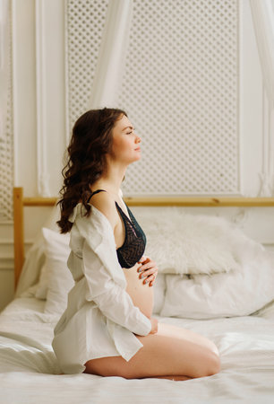 a slender pregnant woman in a black underwea and white shirt on the bedの写真素材