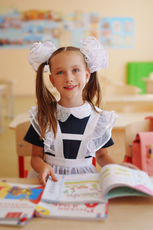 little girl in school uniform sitting at desk with textbooks for first grade.