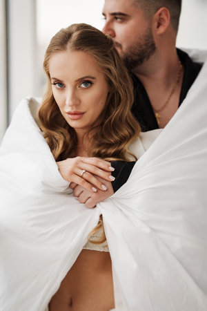 A gentle and romantic man and woman wrapped in a blanket on the windowsill of a large window. Newlyweds. Romance and passion in relationships.の写真素材
