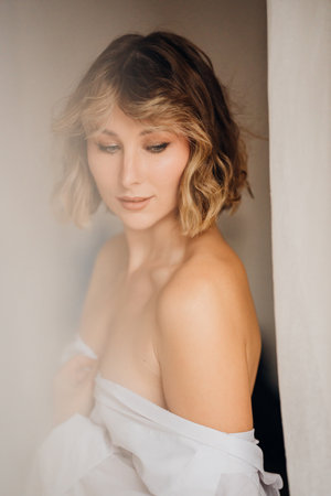 an attractive blonde woman with bare shoulders in a white shirt by the curtain.の写真素材