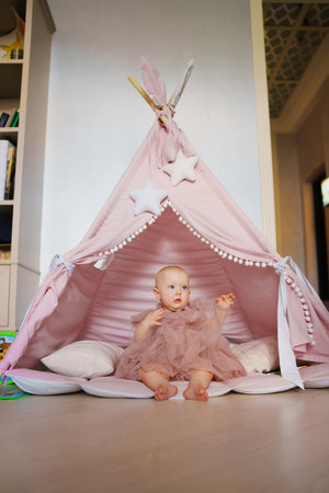 Cute baby girl in pink dress sitting in teepee tent for childrens room.