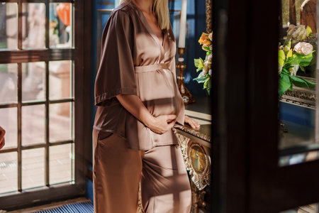 without a face. Pregnant woman in a beige silk suit by the window. The concept of comfortable and beautiful maternity clothesの写真素材