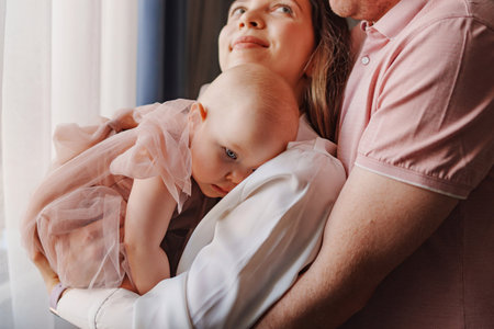 Happy and gentle parents hold baby daughter in pink dress in their arms.