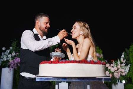 Bride and groom eat a large round cake decorated with berries in the night.