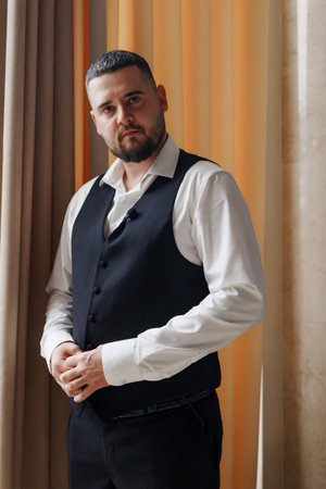 A young man in a vest and a white shirt by the window.
