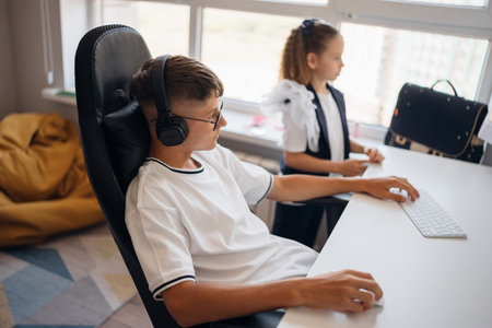Young boy with headphones working at a desk while a girl observes nearbyの写真素材