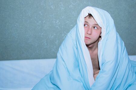 young guy sitting on a bed wrapped in a blue blanket.の写真素材