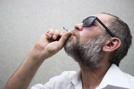 A man in a shirt with a beard in glasses smokes a cigaretteの写真素材