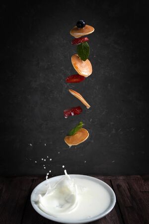 flying tiny pancakes on a dark background, small pancakes with strawberries, mint and blueberries fly out of a plate with milk, flying foodの写真素材
