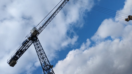 Construction Crane Viewed from Below. Low-angle view of a construction crane against bright blue sky with scattered clouds, emphasizing scale and industrial structure.の写真素材