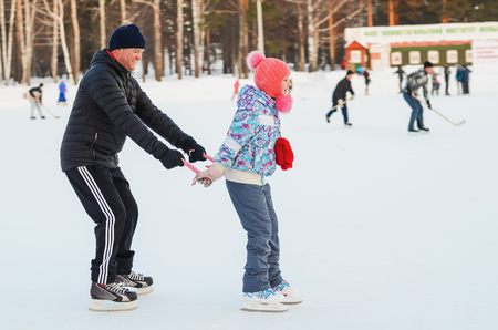 Daddy and daughter skate on the rink under the open sky on a winter eveningの写真素材